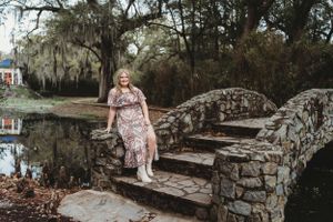 Woman in floral dress stands on stone bridge with moss-draped trees in background. Senior Portrait Photography