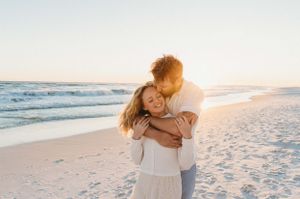 A couple embraces on a sandy beach at sunset while gentle waves roll in along the shoreline.