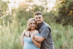 A couple embraces in a sunlit field wearing casual attire during a Maternity photo session.