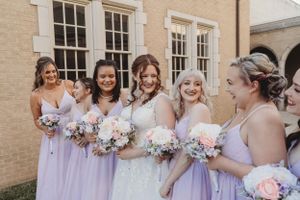 Wedding party wearing light purple bridesmaid dresses hold pastel bouquets while sharing joyful moment outside brick building.