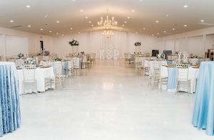 Elegant white wedding reception ballroom with crystal chandelier, blue curtain accents and chiavari chairs arranged at tables.
