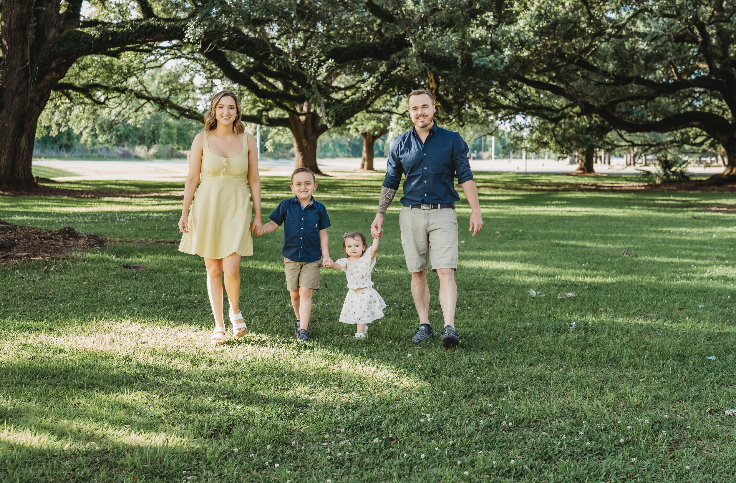 Family of four walking hand-in-hand on green grass under large oak trees in a sunny park.