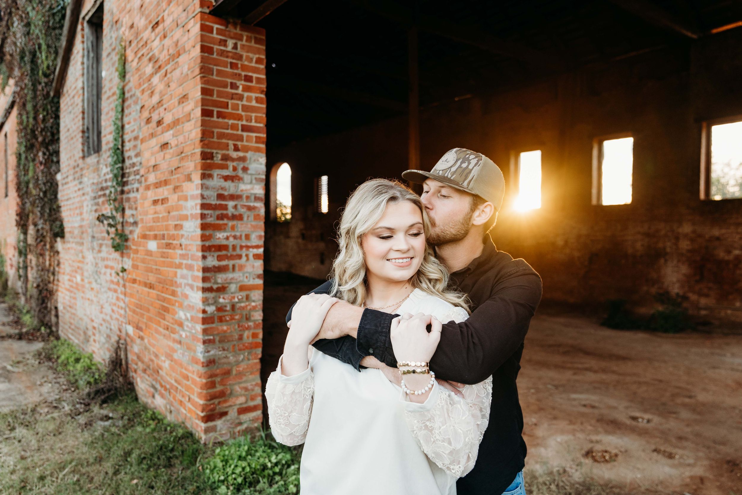 A couple sharing a tender moment against a rustic brick wall with golden sunlight streaming through windows.