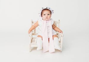 Baby girl in white dress and bonnet sitting in a small decorative white chair on a white background.