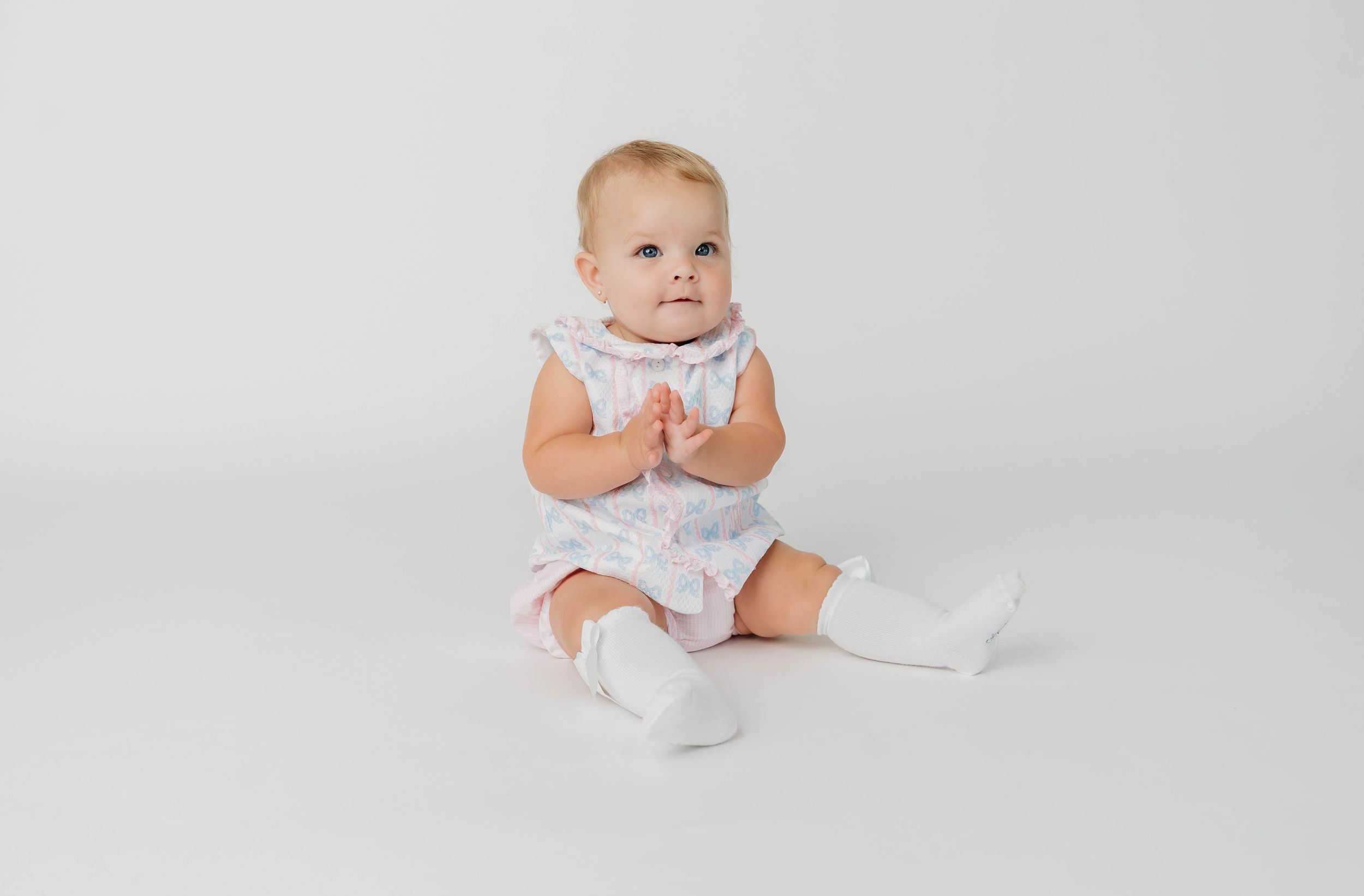 Baby girl sitting in a white floral ruffle dress with white socks on a light gray background.