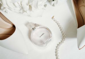 Elegant flatlay composition with Chanel perfume bottle, pearl necklace, nude heels and white floral details.