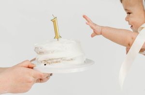 Baby reaching for a white frosted first birthday cake with a gold number one candle on a white plate.