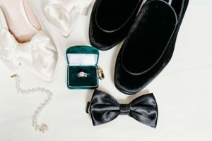 Wedding accessories including black bow tie, dress shoes, engagement ring in green box, and pearl necklace on white background.