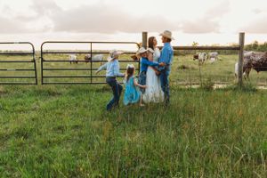 People dance and play in a rustic farm field with metal gates and horses in the background.