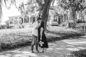 A couple in vintage clothing shares an intimate moment under a Spanish moss-draped tree in front of historic houses.