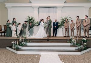 Wedding ceremony in progress at an elegant white church altar decorated with greenery and floral arrangements.