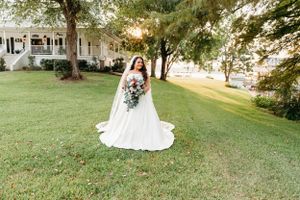 A bride in a white gown holding a bouquet poses on a sunlit lawn near a historic house.