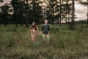 A couple stands among tall grass in a pine forest during sunset.
