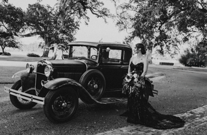 A vintage 1930s Ford car parked outdoors with dark-clad figures beside it.