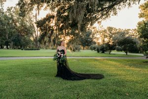 A person in a flowing black dress poses on a sunlit lawn with Spanish moss trees in the background.