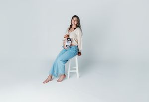 Woman sitting on white stool in light blue flared jeans and beige top, holding a small item, against gray background.