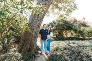 A couple stands beneath a large tree on a garden path during autumn, surrounded by lush foliage.