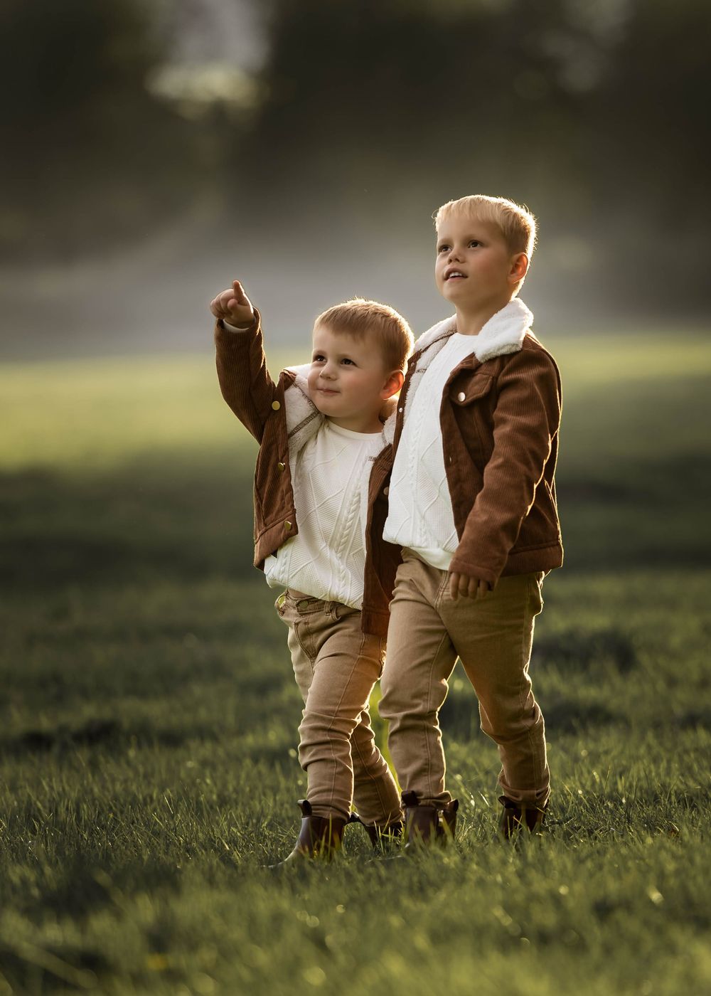 Two children in matching brown jackets and khaki pants stand together in a sunlit grassy field.