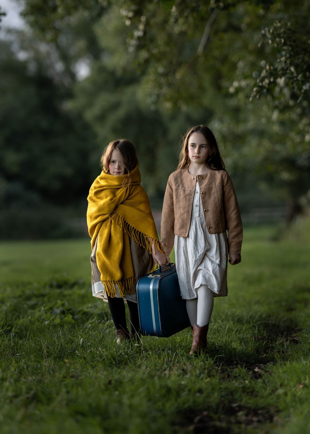 Two people walking through a park, one wearing a yellow shawl and carrying a blue suitcase.