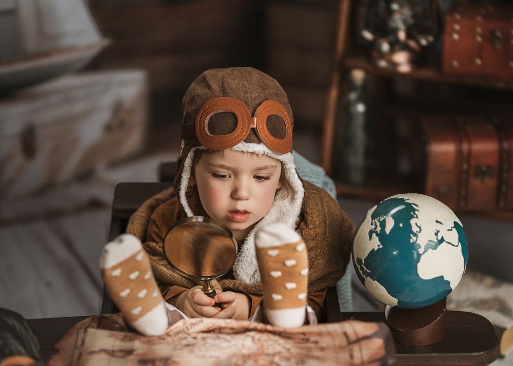 Child in vintage aviator hat studies books and maps with a blue globe on a rustic wooden table.