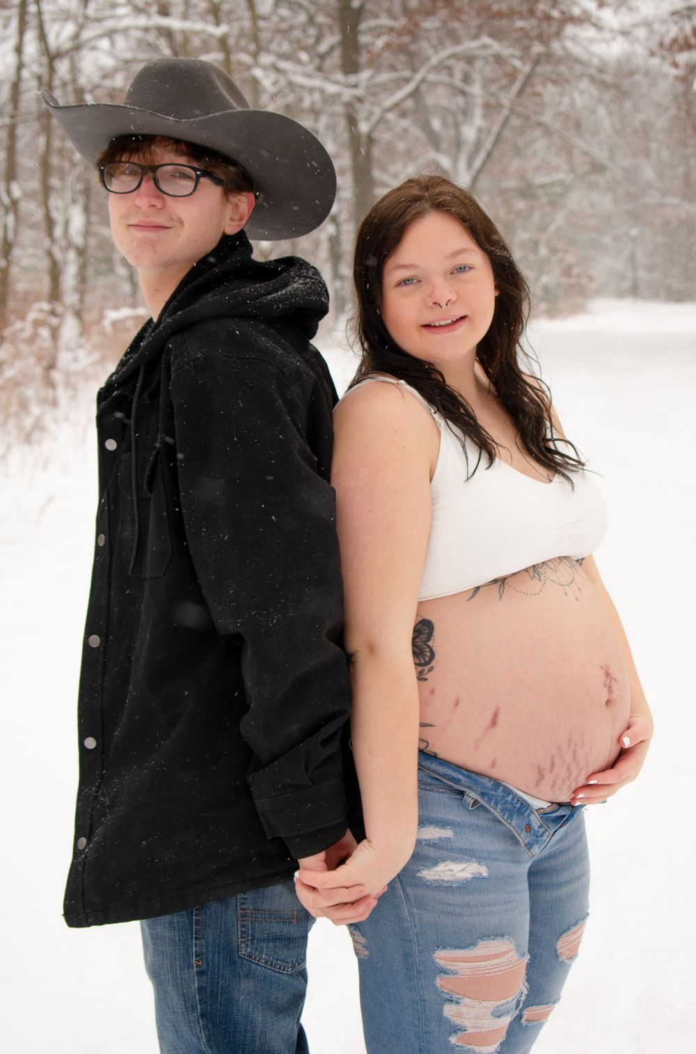 Pregnant mother to be posed back to back with partner, both holding hands and smiling at camera in a Wisconsin woodsy snowy scene