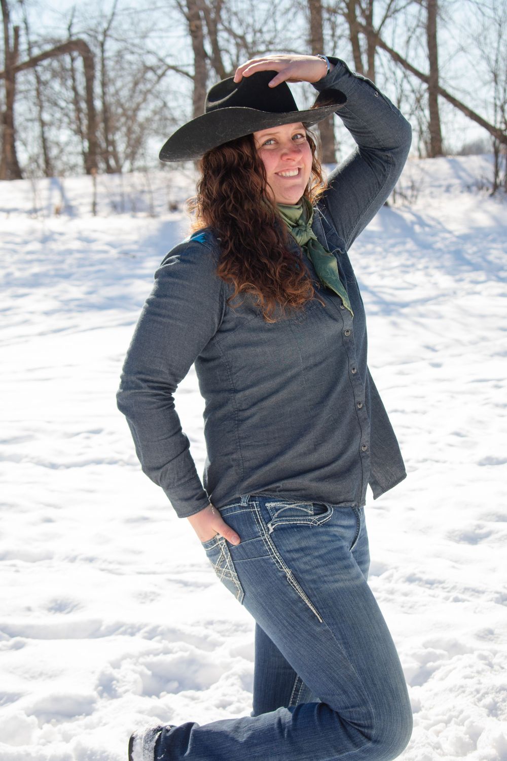 Cowgirl with hand on hat and hand in pocket, smiling at camera in a Wisconsin winter scene