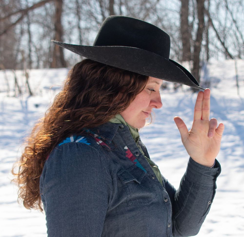 Cowgirl standing sideways with finger gun up to her hat, eyes closed in a Wisconsin winter scene