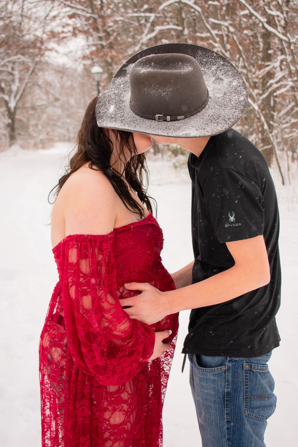 Pregnant mother to be being kissed by partner with cowboy hat blocking their faces in a Wisconsin woodsy winter scene