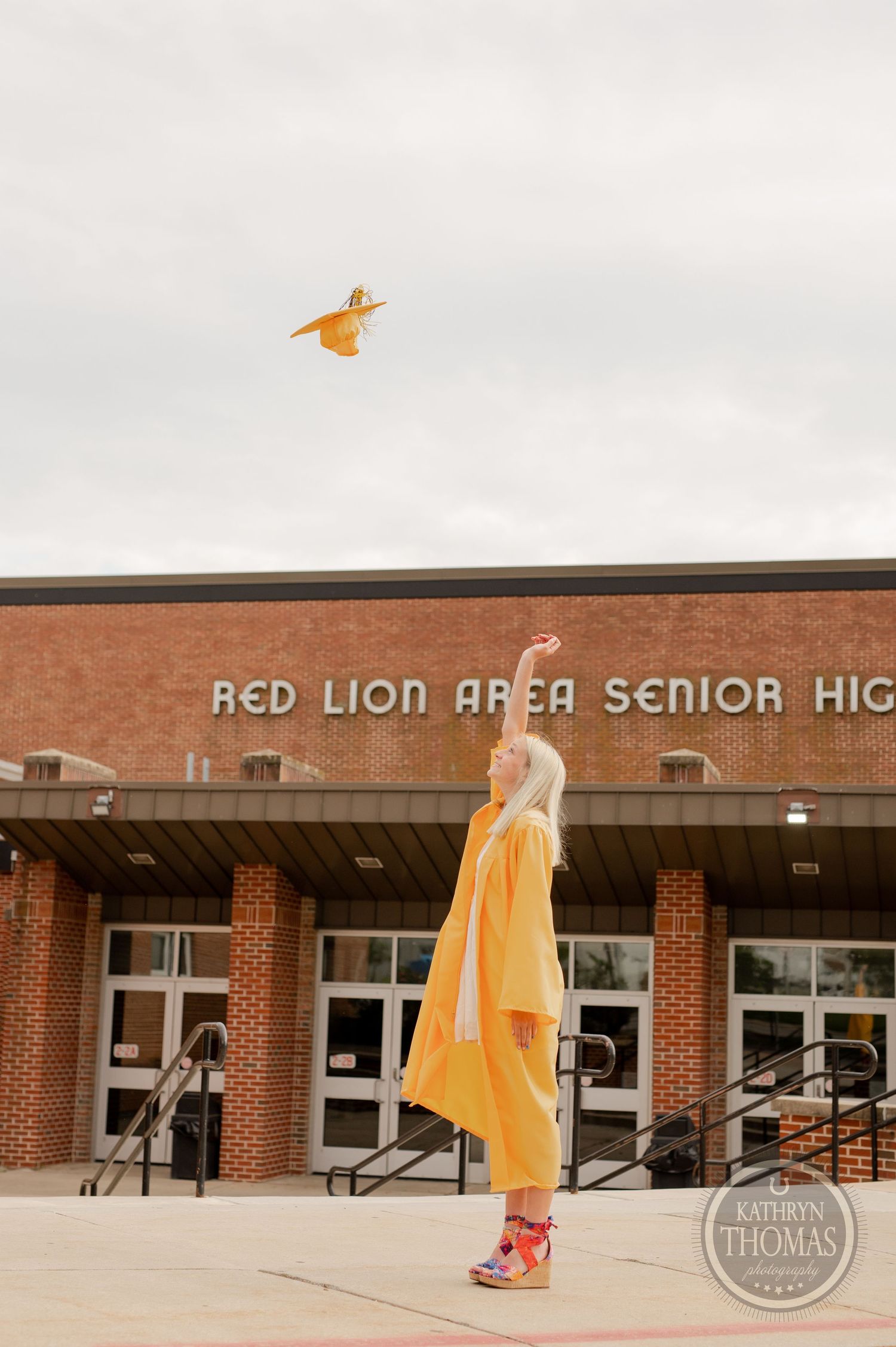 Yellow graduation gown and cap being thrown in celebration outside high school entrance.