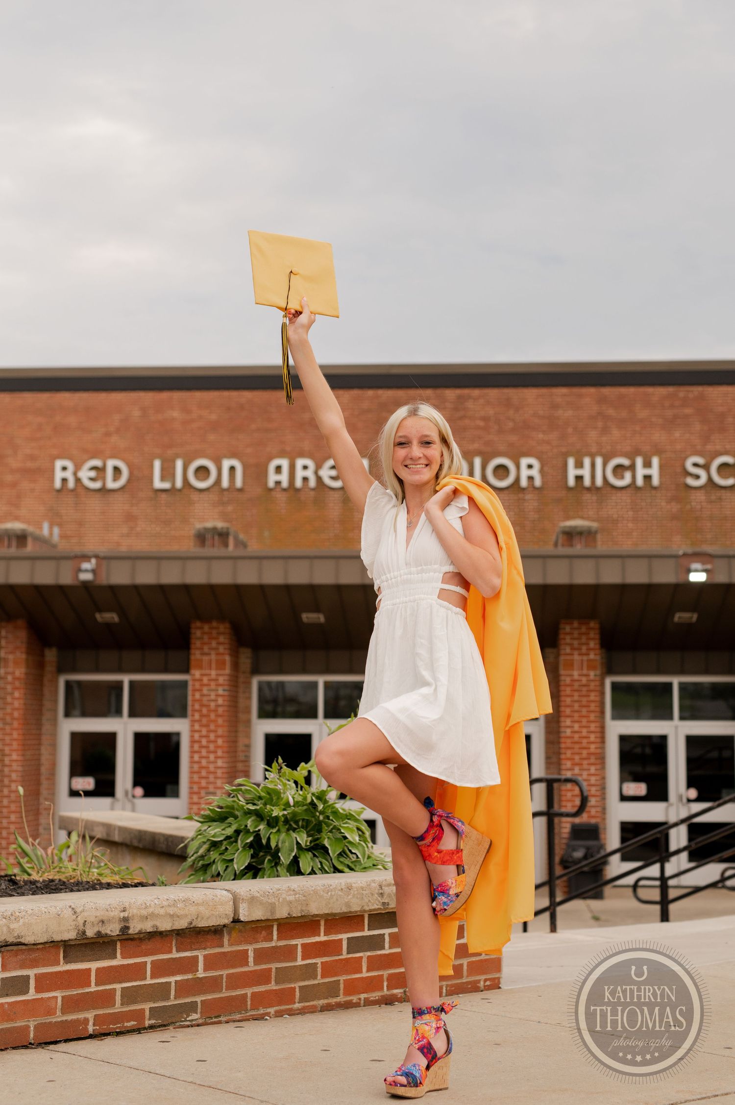 Graduate in white dress raises diploma while posing energetically outside school building.