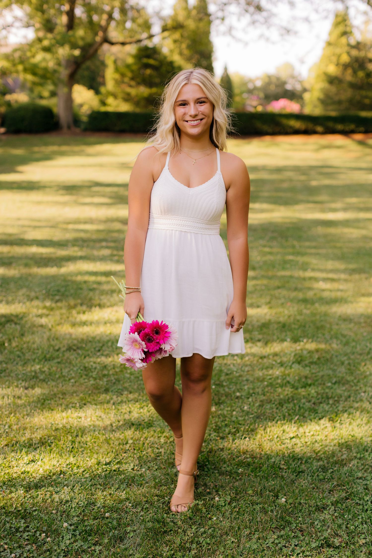 Person in white sundress holds pink flowers while standing on green lawn in summer light.