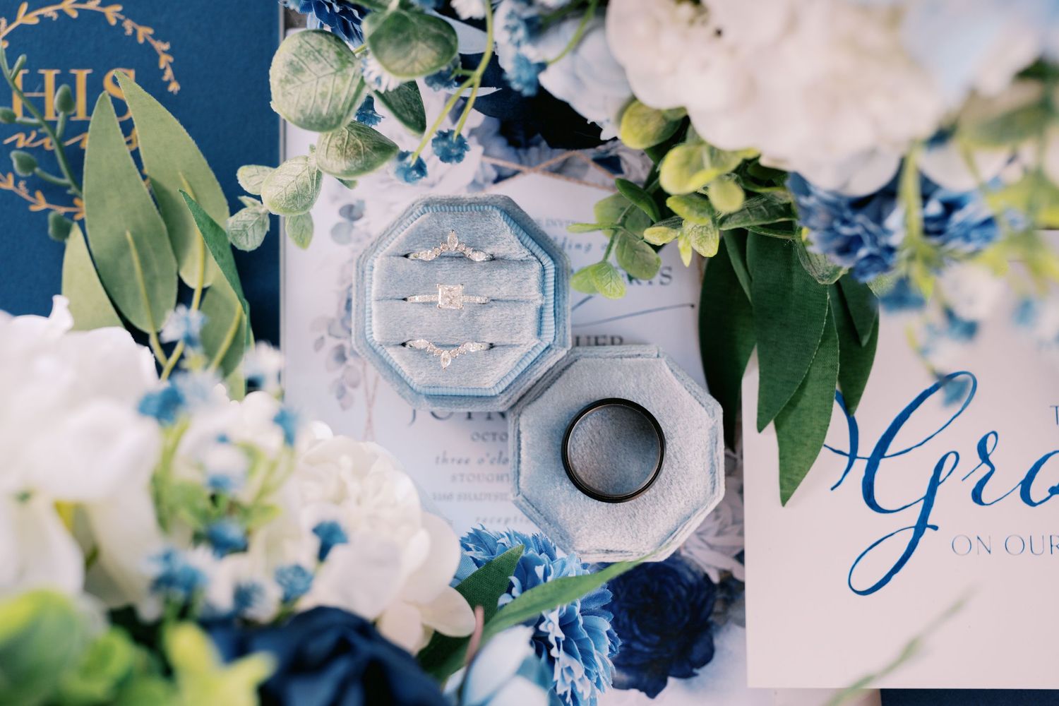 Wedding rings in vintage octagonal box surrounded by white and blue floral arrangements and greenery.