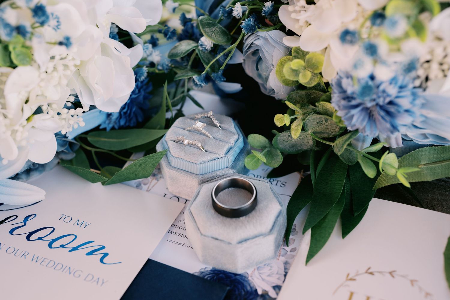 Wedding rings on invitation card surrounded by delicate white and blue floral arrangements with green foliage.