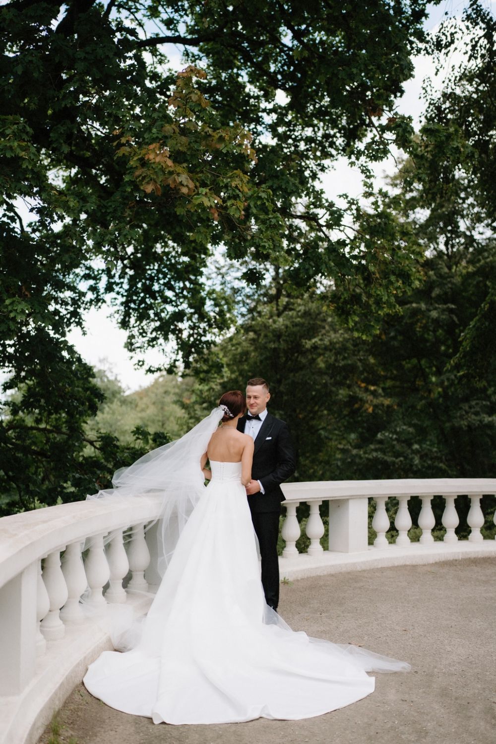 A bride in a white gown stands with her partner on an ornate balcony surrounded by green trees.