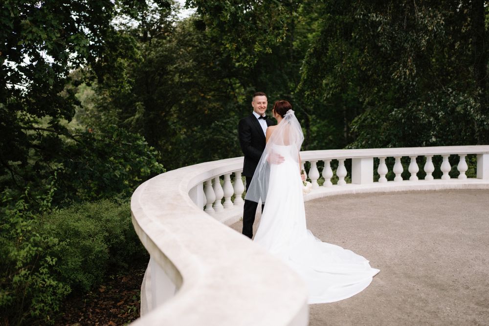 A wedding couple poses elegantly on a white stone terrace with trees in the background.