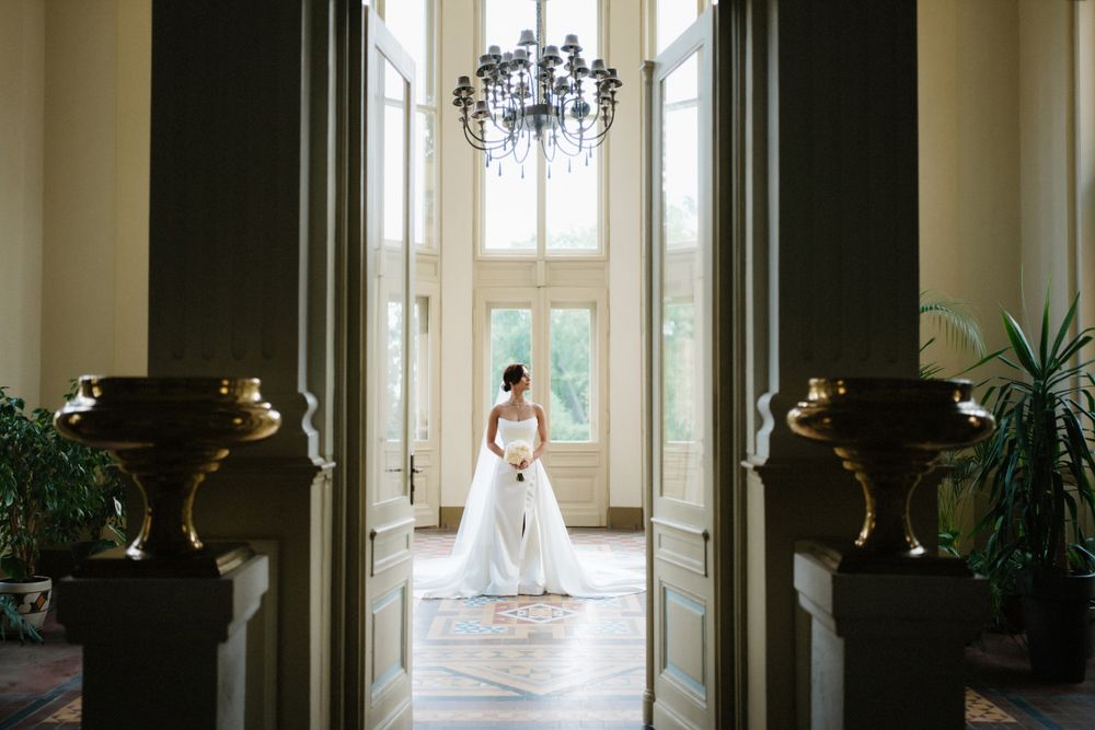 Bride in white gown stands in grand hallway with chandelier and ornate decor.