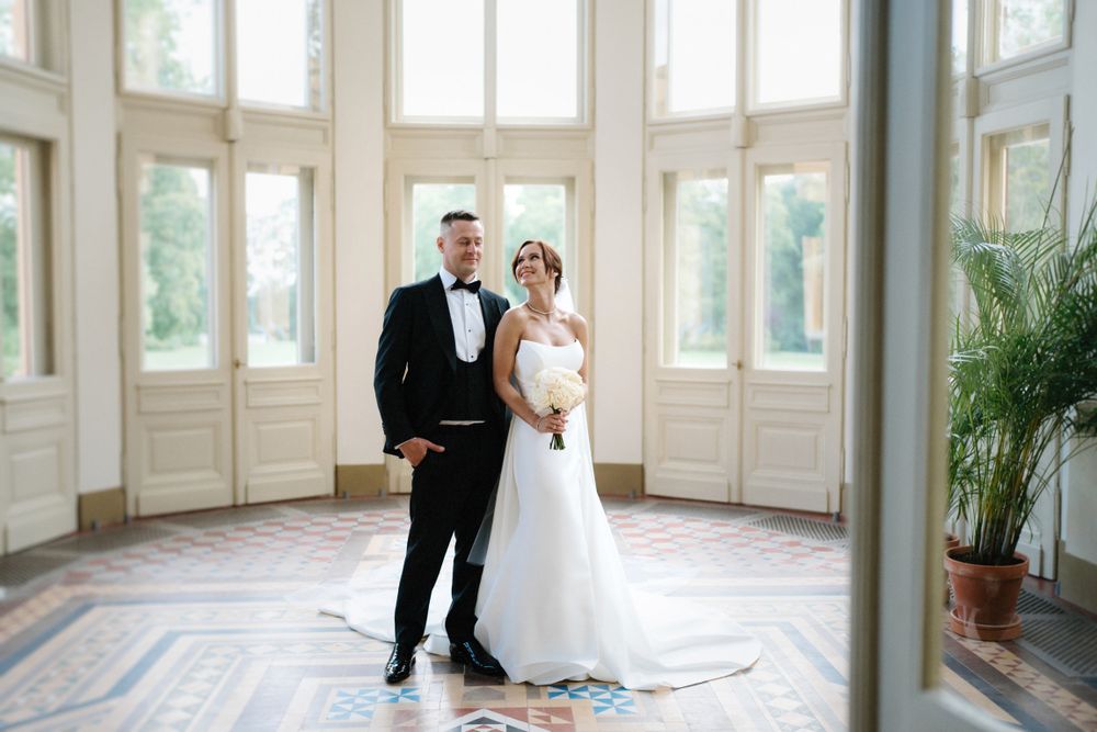 Couple stands in an elegant room with tall windows and ornate white trim.