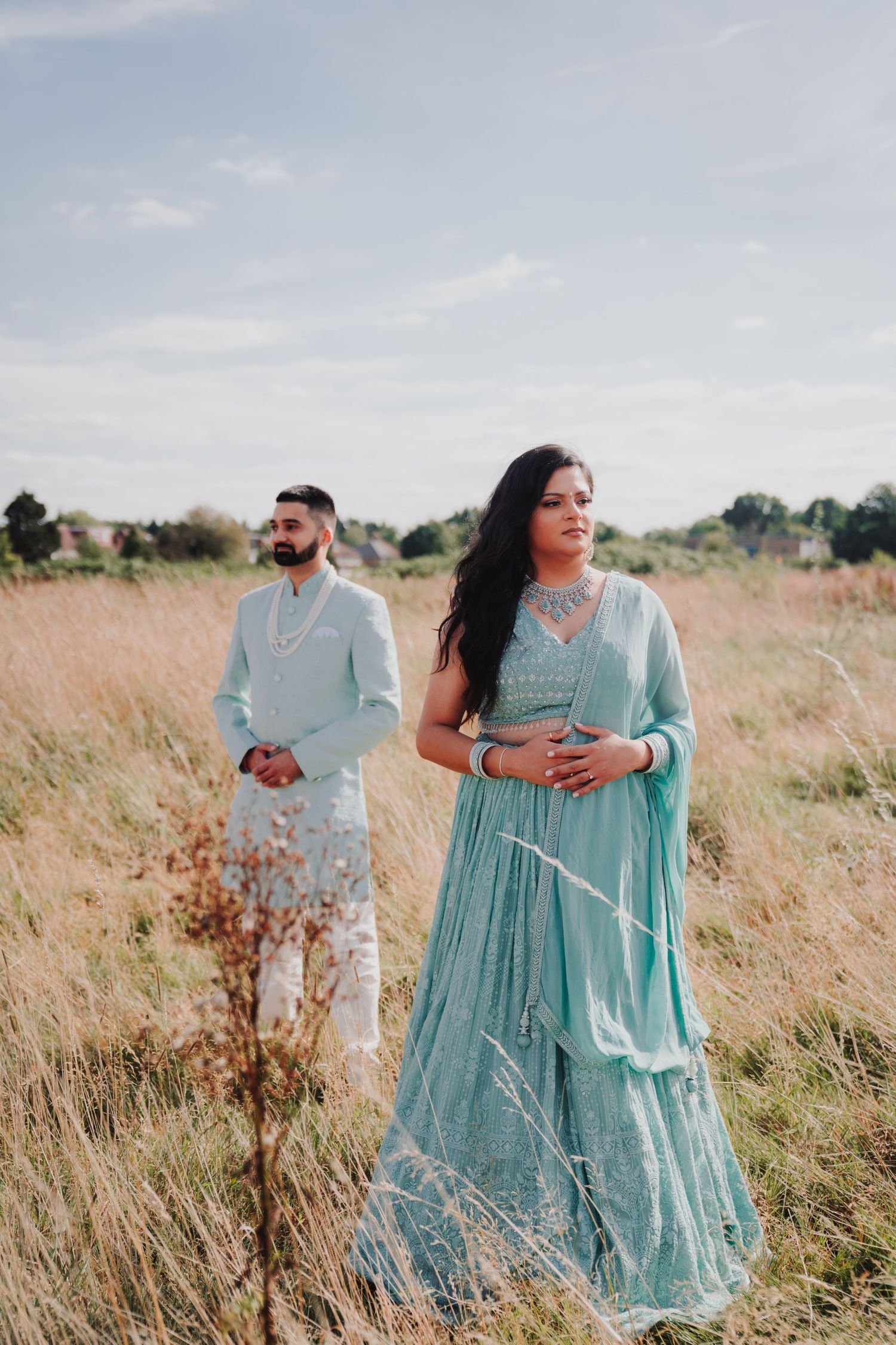 A couple wearing coordinated mint green traditional Indian outfits poses in a golden field at sunset.