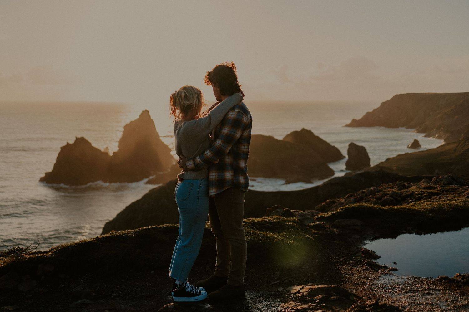 A couple stands together watching the sunset over rocky coastal cliffs and ocean waves.