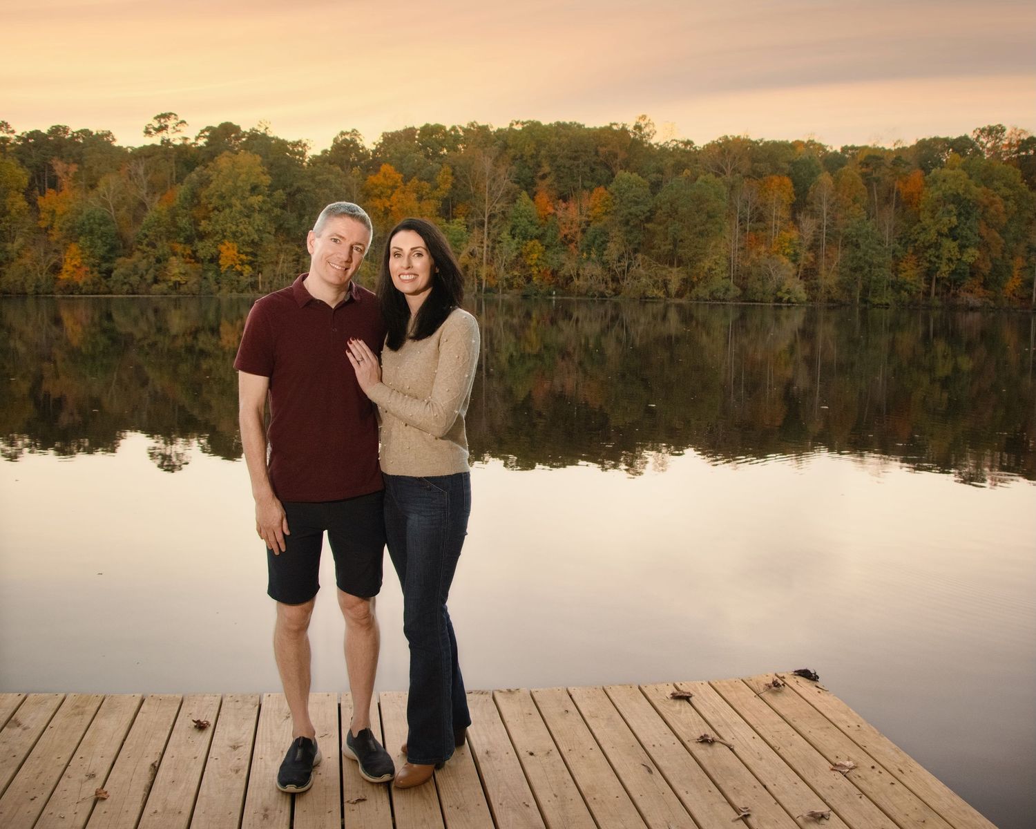 A couple stands together on a wooden dock by a calm lake during autumn with colorful fall foliage reflected in the water.