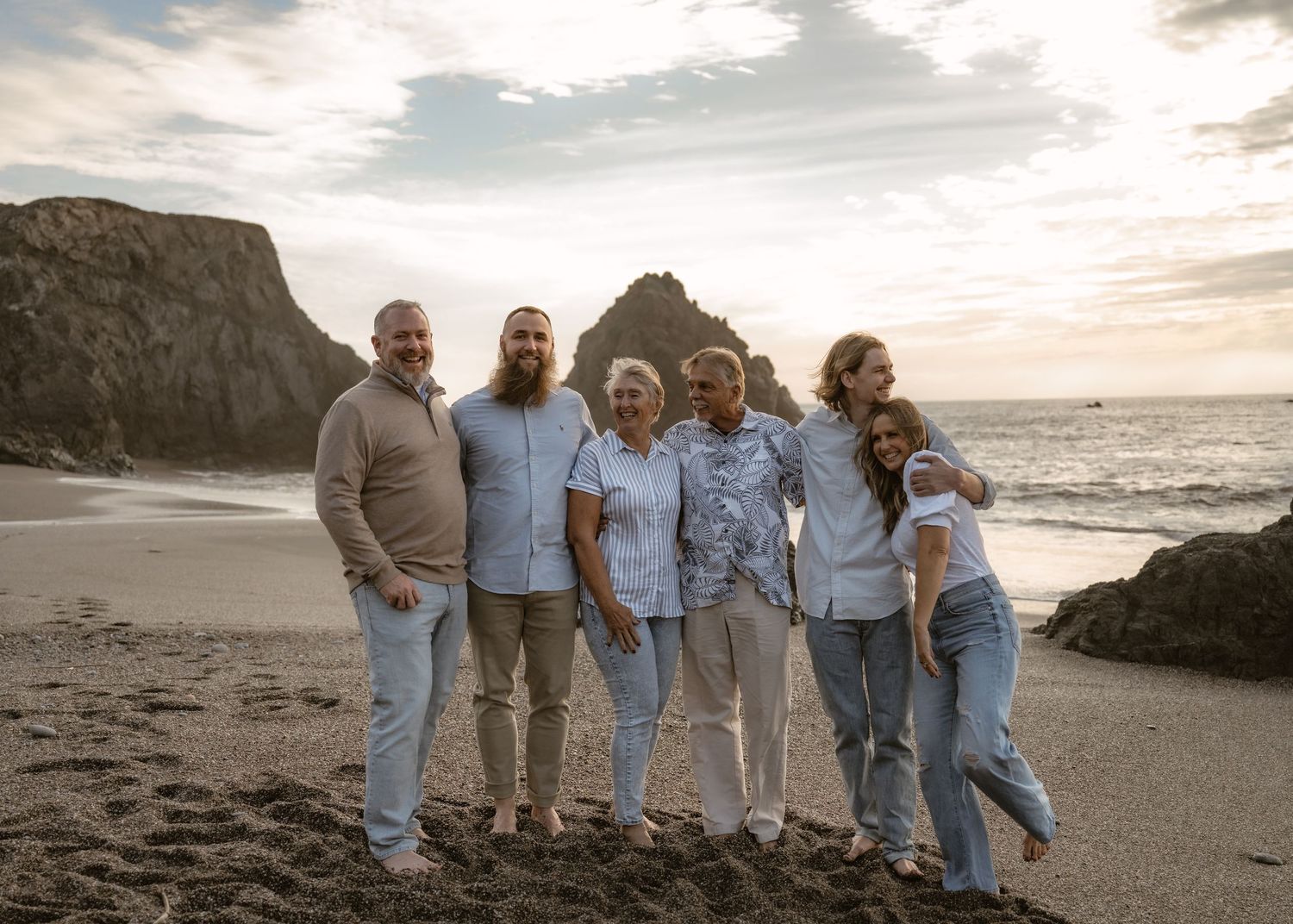 Group wearing coordinated blue and white outfits poses together on a scenic beach at sunset.