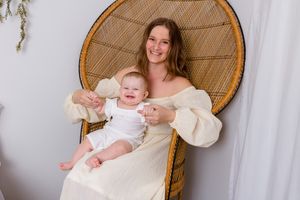 baby girl sits on moms lap both facing front and smiling while sitting on peacock chair