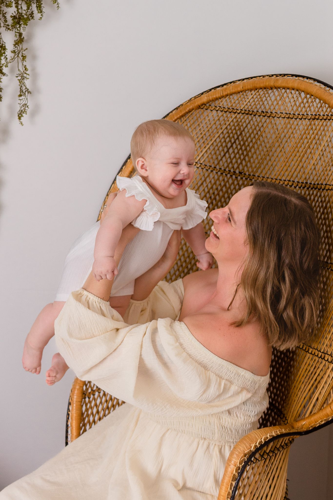 mom holding baby up in the air while sitting on peacock chair against white wall