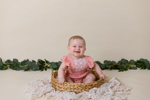 six month old girl in pink outfit sits in wicker basket with cream background