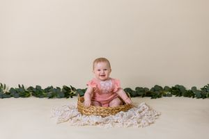 six month old girl in pink outfit sits in wicker basket with cream background
