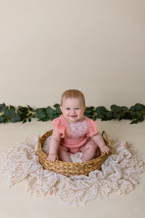 six month old girl in pink outfit sits in wicker basket with cream background