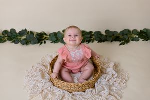 cream background with greenery and six month old girl in pink outfit sits in wicker basket
