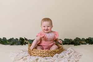 six month old girl in pink outfit sits in wicker basket with cream background