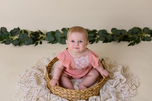six month old girl in pink outfit sits in wicker basket with cream background