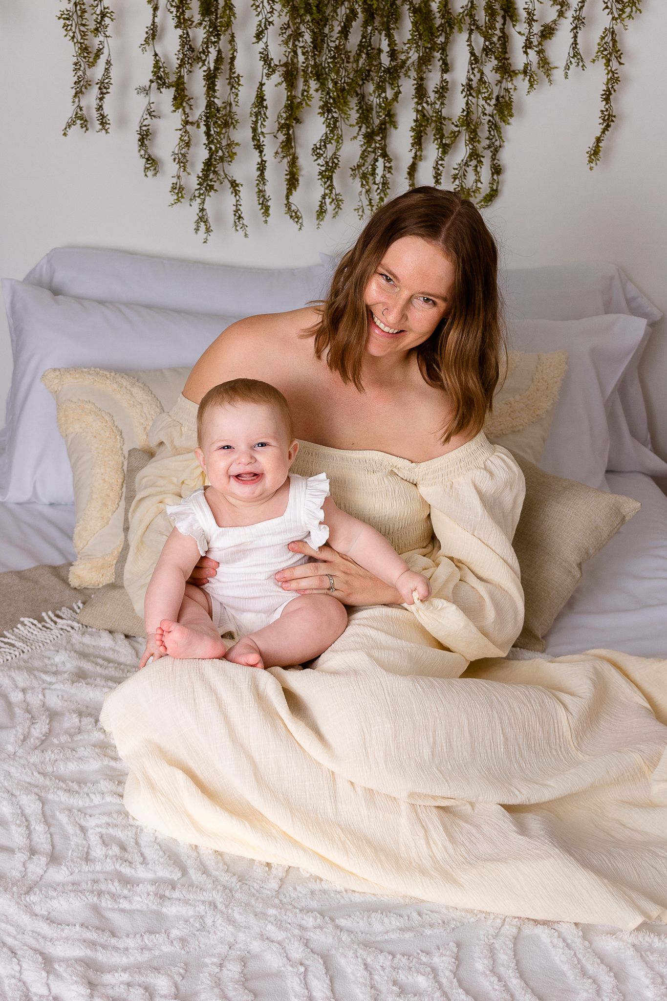 mother sitting on bed holding six month old daughter in lap, both smiling at camera